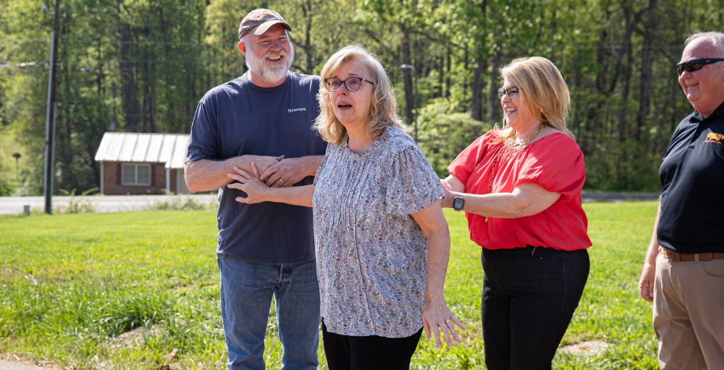 Three adults outdoors, one woman in the center being gently guided by another woman.