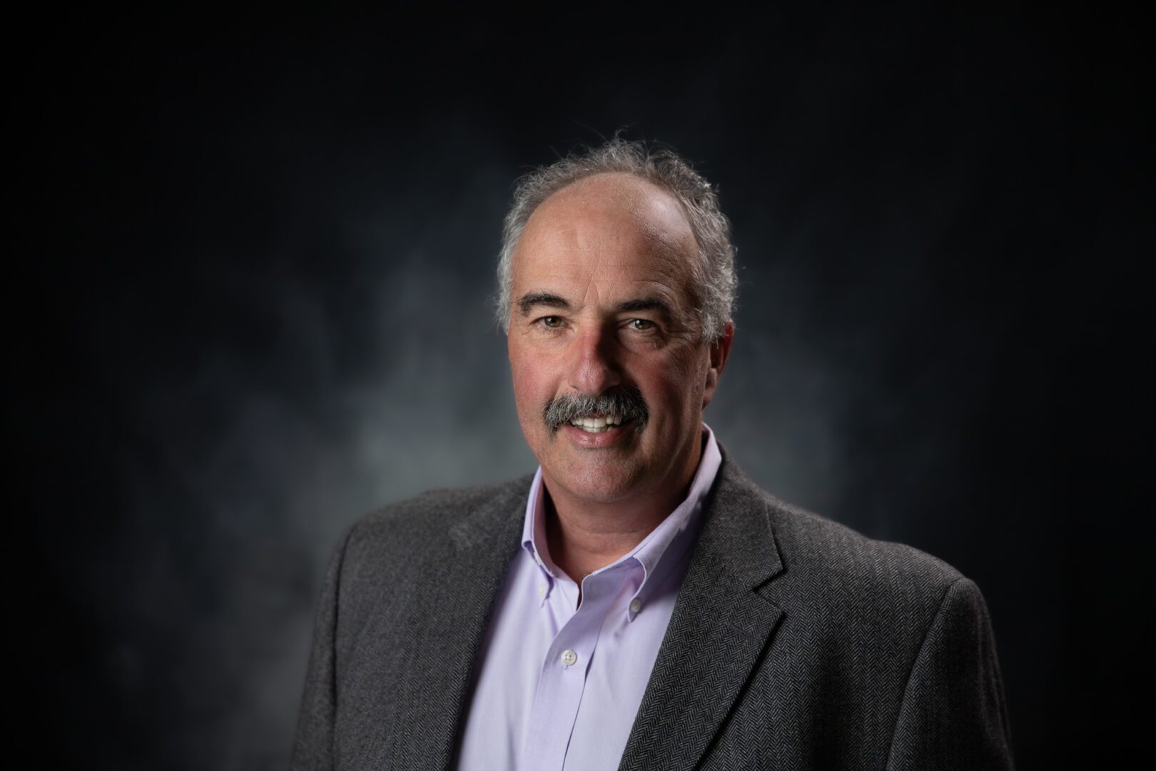 Professional headshot of a man with gray hair and mustache in a suit jacket.