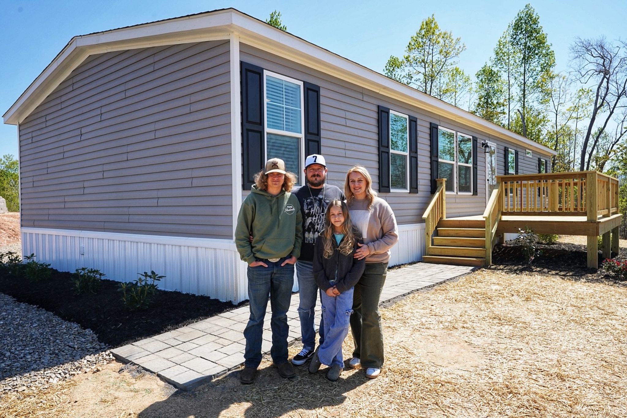Four people stand together smiling outside a house on a sunny day.