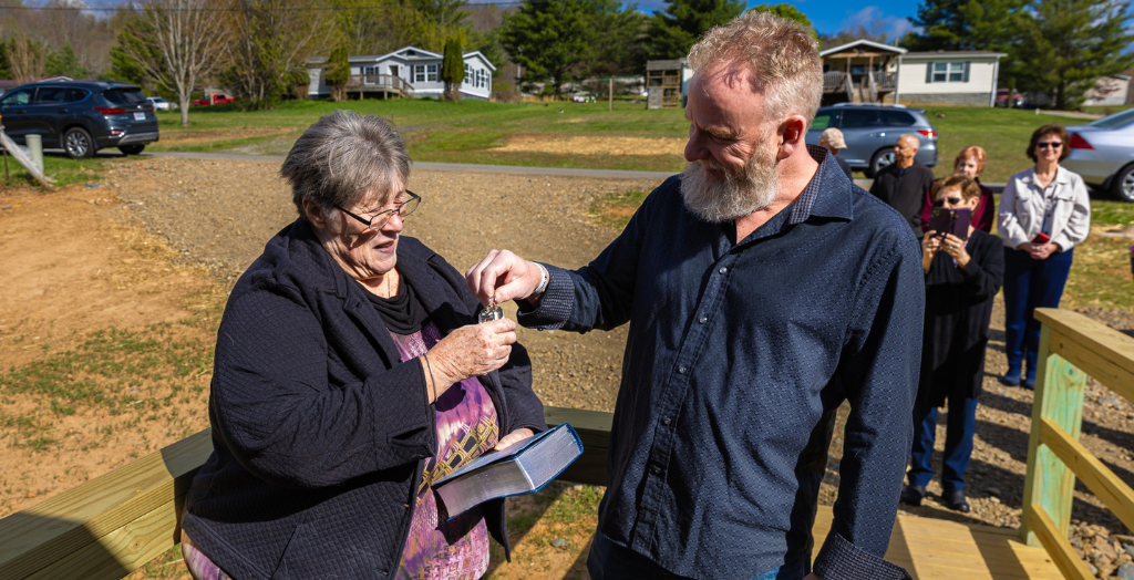An elderly woman and a man exchange keys outdoors on a sunny day.